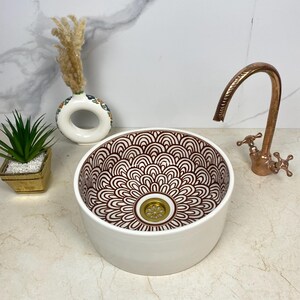 Puede incluir: Lavabo de baño de cerámica blanca con un patrón floral marrón y un desagüe dorado. El lavabo está sobre una encimera blanca con un grifo de cobre.