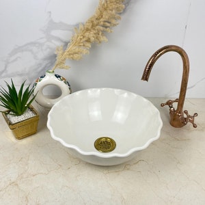 Puede incluir: Lavabo de cerámica blanca con borde acanalado y desagüe de latón. El lavabo está sobre una encimera de mármol blanco con un grifo de cobre y un jarrón decorativo.
