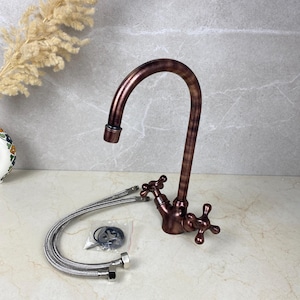 Könnte beinhalten: Ein kupferfarbener Küchenarmatur mit gebogenem Auslauf und kreuzförmigen Griffen. Die Armatur enthält zwei flexible, geflochtene Metallschläuche und einen kleinen Beutel mit Teilen. Die Armatur hat ein Vintage-Aussehen.