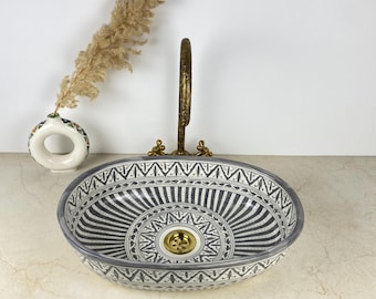Op de afbeelding: Een ovale keramische waskom met een gedetailleerd zwart-wit geometrisch patroon met stralende lijnen en een centrale goudkleurige afvoer. Een hoge, gebogen messing kraan staat achter de waskom, vergezeld door een kleine witte ronde vaas.