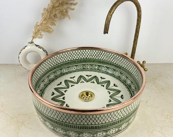 Op de afbeelding: Een ronde keramische waskom met een witte basis en ingewikkelde groene geometrische en florale patronen. De kom heeft een koperkleurige rand en een goudkleurige afvoer. Een sierlijke, gebogen, antiek ogende koperen kraan staat erachter.