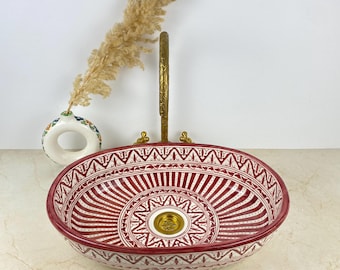 May include: An oval ceramic vessel sink featuring a deep red and white geometric pattern with a central gold-toned drain. Behind the sink stands a tall, slender, antique-style brass faucet with an ornate handle and a small white ceramic vase.