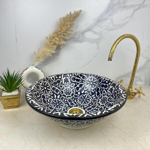以下が含まれることがあります： 白地に黒と白の花柄のセラミック製のシンク。シンクは丸型で、中央に排水口があります。シンクは白いカウンタートップの上に置かれています。