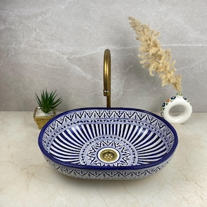May include: An oval, blue and white ceramic bathroom sink with a decorative pattern. The sink has a gold-coloured drain and is set against a neutral background. A gold tap is installed above the sink.