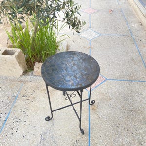 Puede incluir: Una pequeña mesa auxiliar redonda con una superficie de mosaico azul verdoso oscuro. La mesa tiene una estructura de metal negro con adornos decorativos y mide aproximadamente 50 cm de altura.