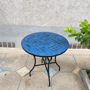 Könnte beinhalten: Ein runder Gartentisch mit einer Mosaikfliesenplatte in Blautönen. Der Tisch hat ein schwarzes Metallgestell mit geschwungenen Beinen. Das Mosaikmuster weist ein geometrisches Design auf. Der Tisch hat einen Durchmesser von ca. 76 cm.