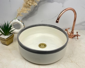 Può includere: Un lavabo in ceramica bianca con un bordo grigio e uno scarico in ottone. Il lavabo è su un piano di lavoro bianco con un rubinetto in rame.