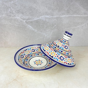 Pode incluir: Um prato de tagine de cerâmica com tampa cónica, apresentando um padrão geométrico vibrante em azul, amarelo, vermelho e branco. O prato tem uma base branca com uma borda azul, ideal para a culinária marroquina.