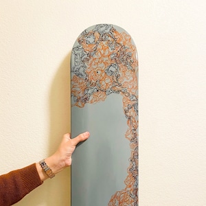 May include: A gray skateboard deck with a black and orange abstract design. The design features a series of swirling lines and shapes.