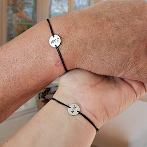 May include: Two matching bracelets. Each has a black cord and a silver-coloured circular charm. The charms display the initials "A" and "C" with a heart symbol. The bracelets are worn on wrists.
