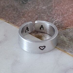 Peut inclure: Une bague réglable en argent avec la lettre "A" estampillée à l'intérieur de la bande et un symbole de cœur à l'extérieur. La bague est présentée sur une surface en marbre.