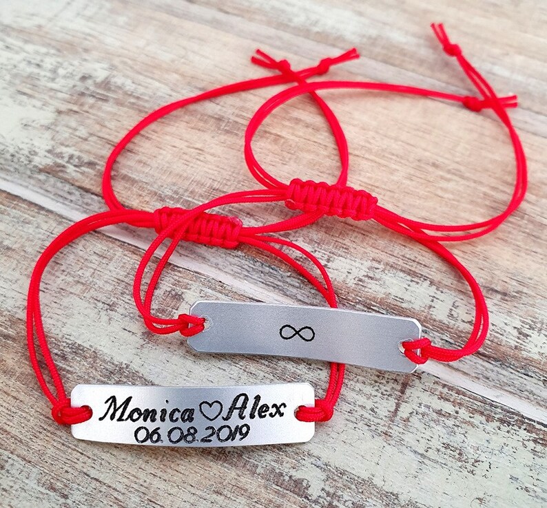 Pu&ograve; includere: Due braccialetti abbinati con cordini rossi e targhette rettangolari argentate. Uno ha il simbolo dell'infinito, l'altro i nomi "Monica" e "Alex" con un cuore e la data "06.08.2019".