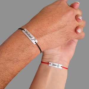 May include: Two matching bracelets with black and red string. The bracelet on the left has a silver tag with the word "Michael" and a key symbol. The bracelet on the right has a silver tag with the word "Jessi" and a padlock symbol.