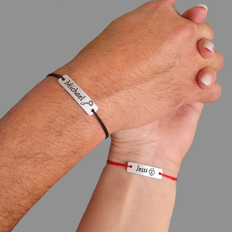 May include: Two matching bracelets with black and red string. The bracelet on the left has a silver tag with the word "Michael" and a key symbol. The bracelet on the right has a silver tag with the word "Jessi" and a padlock symbol.