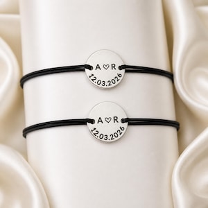 Puede incluir: Dos pulseras a juego con cordones negros y colgantes circulares plateados. Cada colgante tiene las iniciales "A ♡ R" y la fecha "12.03.2026" grabadas. Las pulseras se muestran sobre un fondo blanco y sedoso.