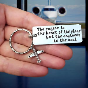 Puede incluir: Llavero plateado con un pequeño dije de avión y una etiqueta rectangular. La etiqueta tiene la cita: "El motor es el corazón del avión, pero el ingeniero es el alma". El fondo muestra una imagen borrosa de un avión a reacción.