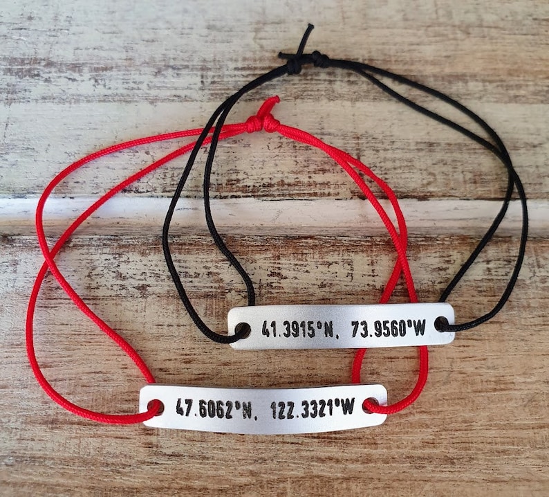 Puede incluir: Dos pulseras personalizadas con coordenadas grabadas en barras de metal plateado. Una pulsera tiene un cord&oacute;n rojo, la otra un cord&oacute;n negro. Las coordenadas est&aacute;n en texto negro. Las pulseras est&aacute;n sobre una superficie de madera clara.