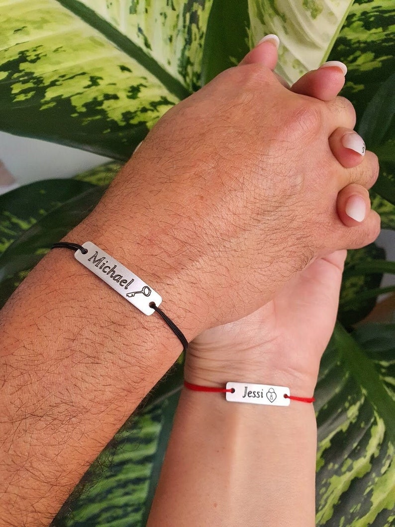 May include: Two matching bracelets with black and red cords. The bracelets have silver metal tags with the names "Michael" and "Jessi" engraved on them. Each tag has a small heart symbol.