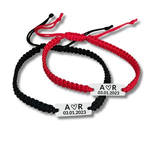 Puede incluir: Dos pulseras de amistad a juego. Una es negra, la otra roja. Cada pulsera tiene una etiqueta rectangular plateada con las iniciales "A♡R" y la fecha "03.01.2023" impresas. Las pulseras están sobre un fondo blanco.