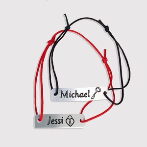 May include: Two matching bracelets with red and black cords. One bracelet has a silver tag with the word "Michael" and a key symbol. The other bracelet has a silver tag with the word "Jessi" and a padlock symbol.