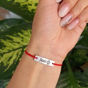 May include: Red string bracelet with a silver charm that says "Jessi" with a small padlock symbol.