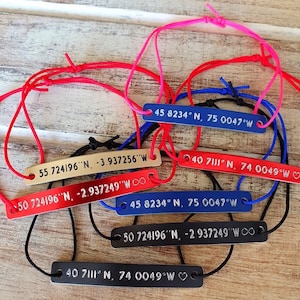 Puede incluir: Varios brazaletes con etiquetas rectangulares de metal y cordones ajustables de varios colores. Cada etiqueta muestra coordenadas en texto blanco. Los cordones son rojos, rosas, azules y negros.