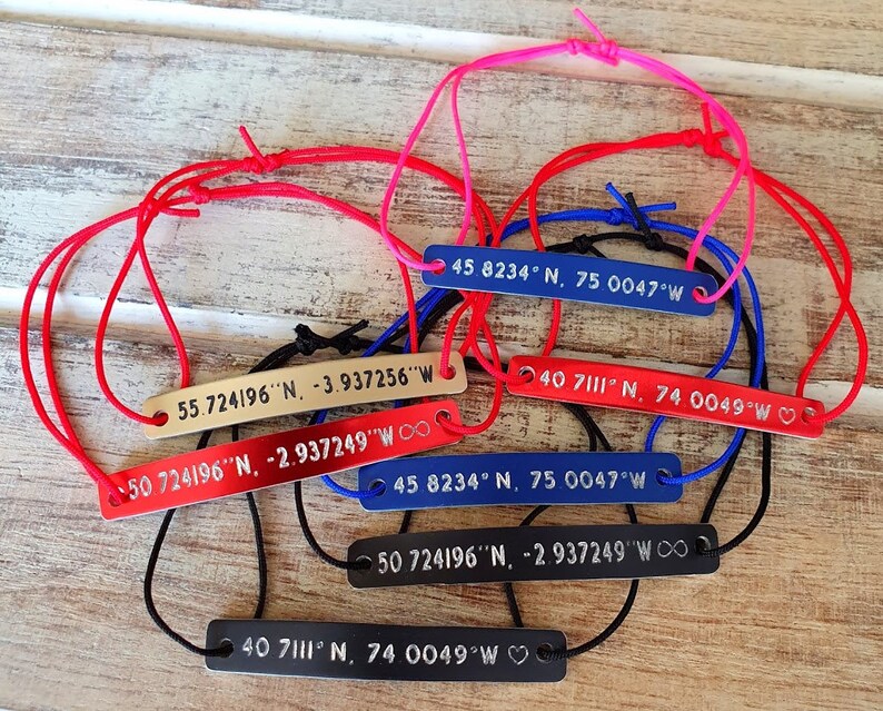 Puede incluir: Varios brazaletes con etiquetas rectangulares de metal y cordones ajustables de varios colores. Cada etiqueta muestra coordenadas en texto blanco. Los cordones son rojos, rosas, azules y negros.