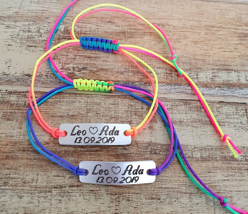 Pu&ograve; includere: Due braccialetti abbinati con cordini neon colorati in rosa, giallo, blu e verde. Ogni braccialetto presenta una placca rettangolare argentata con i nomi "Leo ♡ Ada" e la data "13.09.2019" incisi. I braccialetti sono su una superficie di legno.