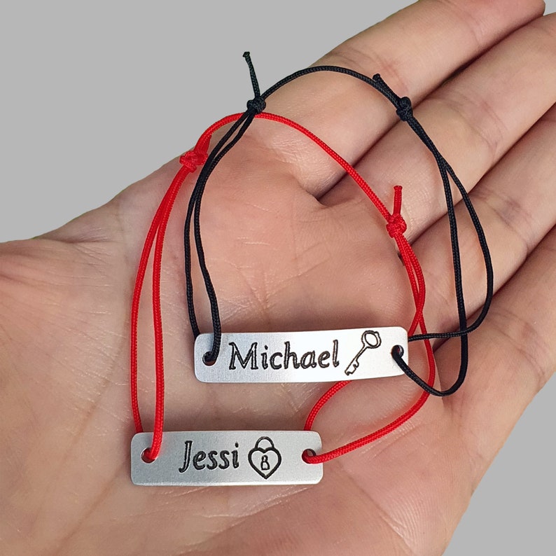 May include: Two matching bracelets with personalized engraved tags. The red bracelet has a tag that says "Jessi" with a heart and padlock symbol. The black bracelet has a tag that says "Michael" with a key symbol.