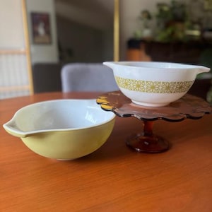 May include: Two vintage Pyrex bowls. One is pale yellow with a white interior. The other is white with a gold geometric pattern. The bowls are on a wooden table, with a brown glass cake stand.