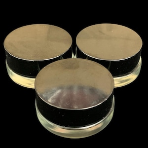 May include: Three small, round, clear glass jars arranged in a triangular formation. Each jar features a reflective, metallic silver lid with a smooth, flat top and a black band around the side, creating a uniform and clean appearance.