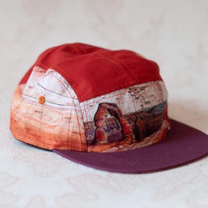 May include: A five-panel cap featuring a solid red crown top, a flat dark purple brim, and a side panel printed with a detailed landscape illustration of a natural rock arch formation in a desert setting with orange and brown earth tones.