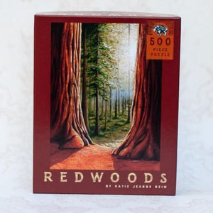 May include: A boxed 500-piece puzzle featuring a forest scene with towering redwood trees. The box is a deep red with the word "REDWOODS" in gold lettering. The puzzle is by Katie Jeanne Reim.