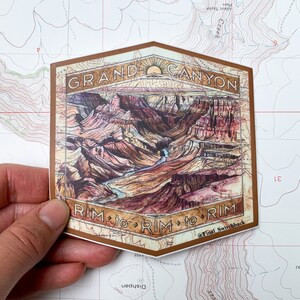 May include: A brown and tan sticker with a vintage illustration of the Grand Canyon. The sticker has the text "GRAND CANYON" at the top and "RIM to RIM to RIM @Final Switchback" at the bottom.