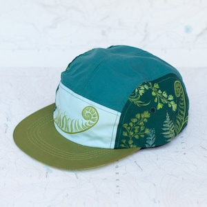 May include: A five-panel cap in shades of teal, green, and olive. The cap features a light blue panel with a fern design and a dark green panel with a botanical print. The brim is olive green.