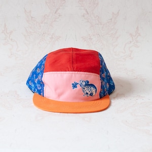 May include: A five-panel cap featuring a red top panel, a light pink front panel with a blue graphic of a small rodent holding a leaf, blue side panels with a floral pattern, and a solid orange flat brim. The cap has a structured crown design.