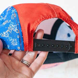 May include: A five-panel cap featuring a vibrant blue fabric with a white and orange floral pattern on the side panels, contrasted by a solid red center panel. The back includes a black plastic snapback closure for adjustable sizing and fit.