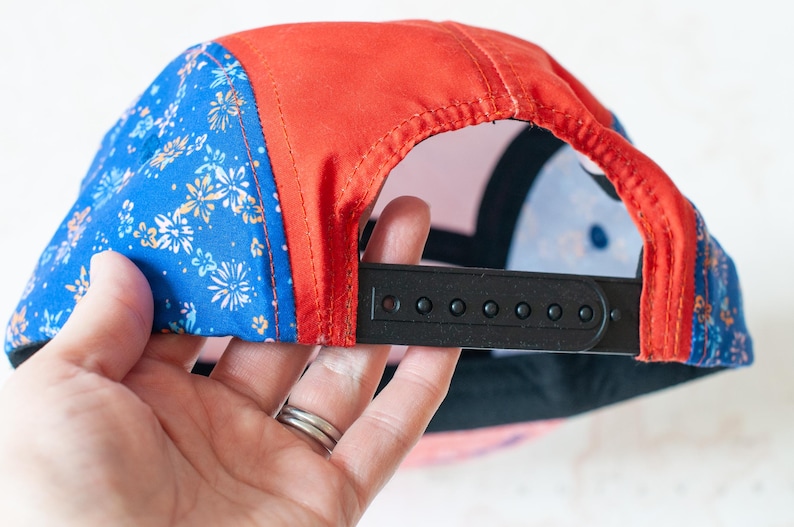May include: A five-panel cap featuring a vibrant blue fabric with a white and orange floral pattern on the side panels, contrasted by a solid red center panel. The back includes a black plastic snapback closure for adjustable sizing and fit.