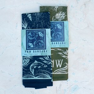 May include: Two folded bandanas in a blue and green colour scheme. The bandanas feature a nature-inspired design with a salmon fish, pine cones, and floral elements. The text "PNW Bandana" is printed on the blue bandana. The text "Bandana" is printed on the green bandana.