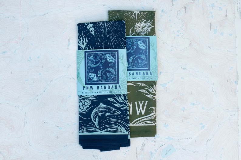 May include: Two folded bandanas in a blue and green colour scheme. The bandanas feature a nature-inspired design with a salmon fish, pine cones, and floral elements. The text "PNW Bandana" is printed on the blue bandana. The text "Bandana" is printed on the green bandana.