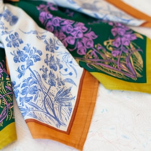 May include: A set of three square scarves featuring botanical illustrations. One scarf is dark green with purple flowers and a mustard border, another is white with blue flowers and an orange border, and the third is green with purple floral prints.