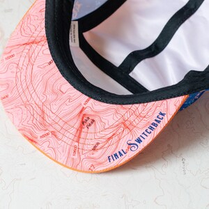 May include: A five-panel cap with a pink brim featuring a topographic map print, the text Pika Peak, and the text FINAL SWITCHBACK in blue. The interior has black seam tape and a white lining, with a blue patterned exterior fabric visible.