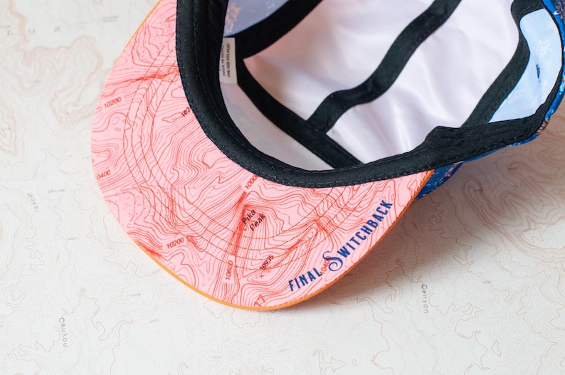 May include: A five-panel cap with a pink brim featuring a topographic map print, the text Pika Peak, and the text FINAL SWITCHBACK in blue. The interior has black seam tape and a white lining, with a blue patterned exterior fabric visible.