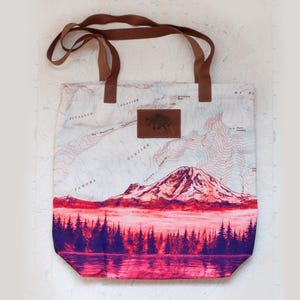 May include: A canvas tote bag featuring a mountain landscape design. The bag has brown leather handles and a leather patch with an animal silhouette. The bottom of the bag has a purple and pink forest scene, with a mountain in the center.