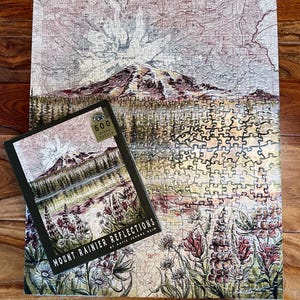 May include: A completed 500-piece jigsaw puzzle featuring a landscape of Mount Rainier and a matching box. The puzzle and box art depict a mountain scene with a lake, flowers, and the text "Mount Rainier Reflections". The puzzle is on a wooden surface.