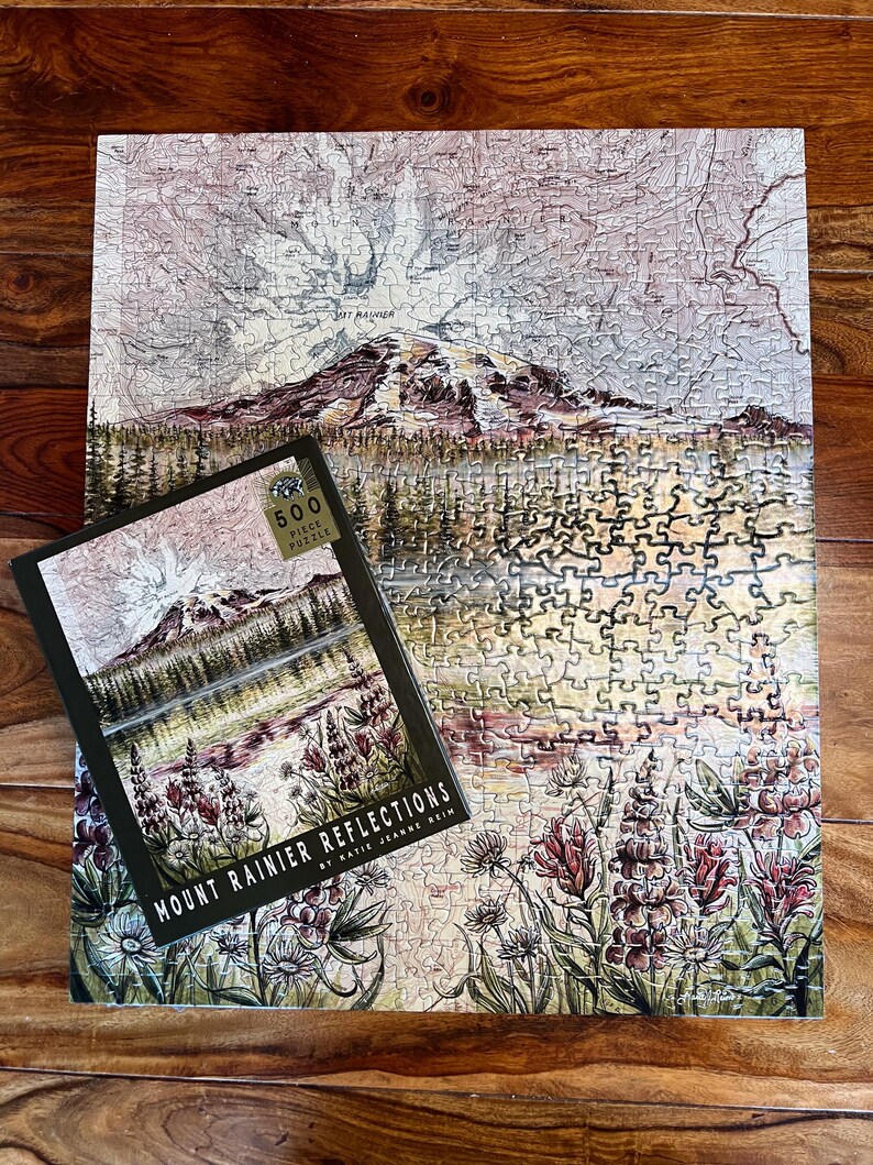 May include: A completed 500-piece jigsaw puzzle featuring a landscape of Mount Rainier and a matching box. The puzzle and box art depict a mountain scene with a lake, flowers, and the text "Mount Rainier Reflections". The puzzle is on a wooden surface.