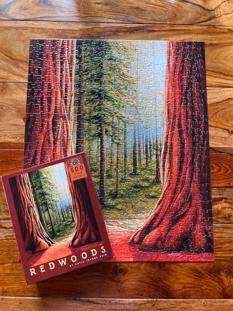 May include: A completed 500-piece jigsaw puzzle depicting a redwood forest scene, with towering trees and a path leading into the distance. The puzzle is next to its box, which also shows the forest scene and the word "Redwoods".