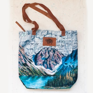 May include: A canvas tote bag featuring a mountain landscape design with a lake and forest. The bag has brown leather handles and a leather patch with a bison graphic. The background has a map design.