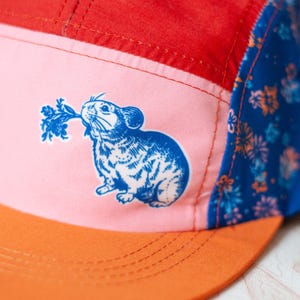 May include: A five-panel cap featuring a red top panel, a light pink front panel with a blue graphic of a rodent holding a leafy plant, and a blue side panel with a floral pattern. The cap has a solid orange flat brim with visible stitching.
