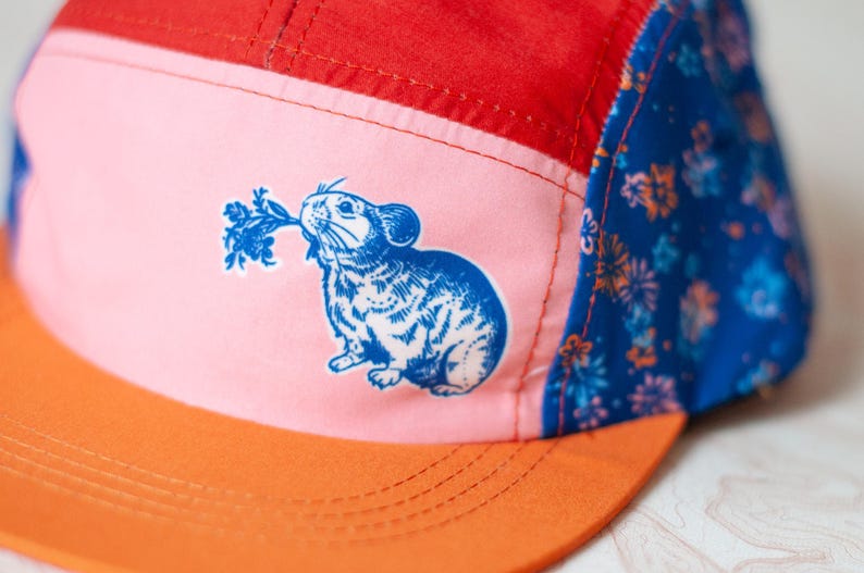 May include: A five-panel cap featuring a red top panel, a light pink front panel with a blue graphic of a rodent holding a leafy plant, and a blue side panel with a floral pattern. The cap has a solid orange flat brim with visible stitching.